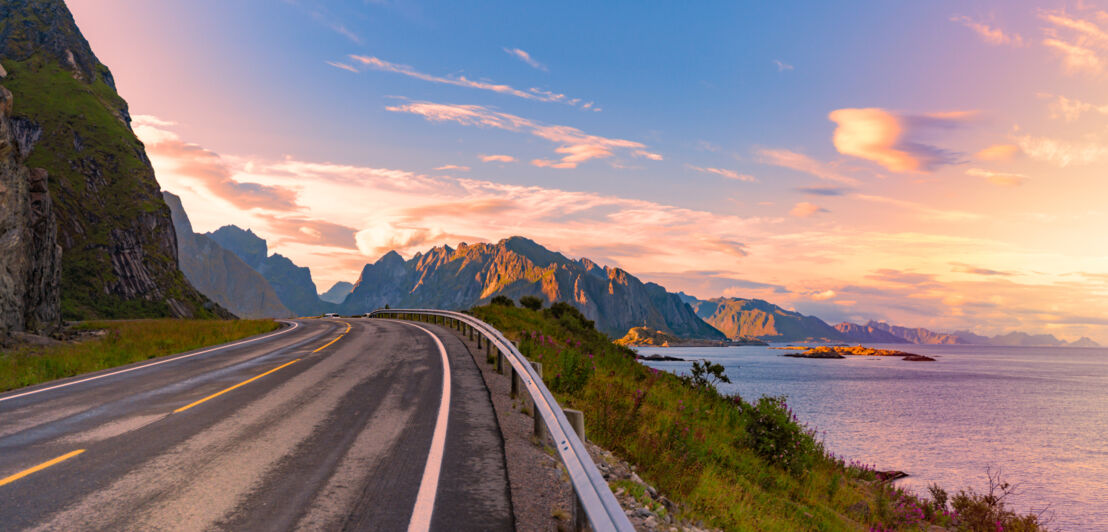 Panorama einer Straße bei Sonnenuntergang auf den norwegischen Lofoten, die am Meer entlangführt. Im Hintergrund Berge.