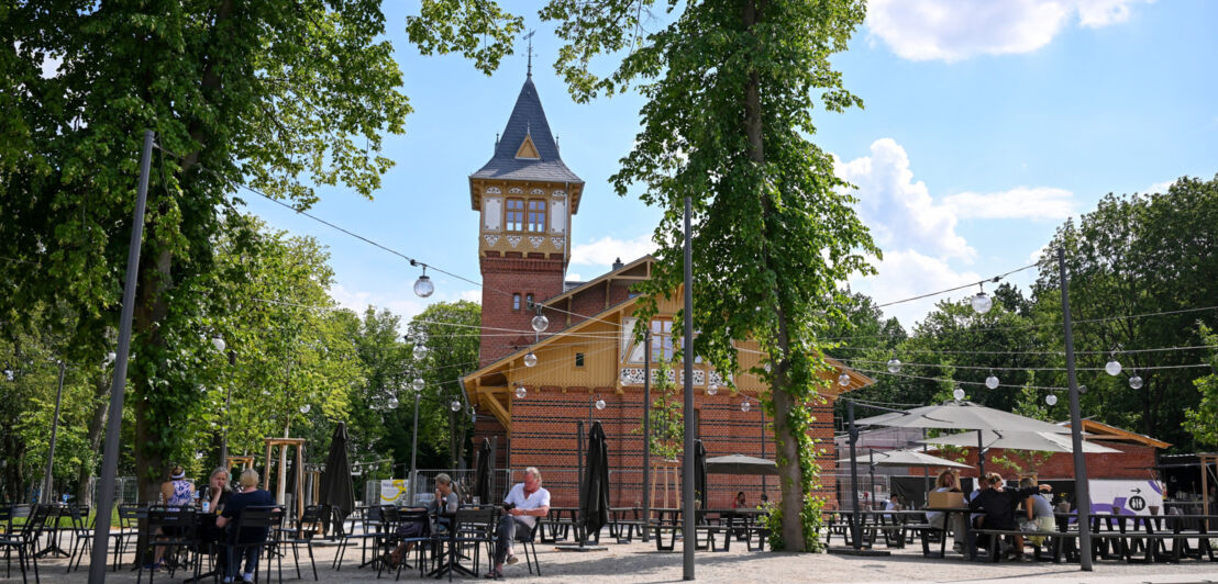 Personen in einem Biergarten vor einem Backsteinhaus mit Turm.