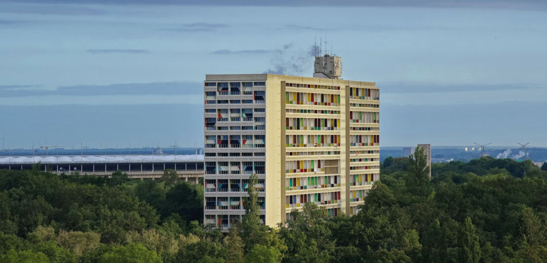 Luftaufnahme eines von Wald umgebenen Hochhauses im Corbusier-Stil mit gemusterter Fassade vor einem Stadion.