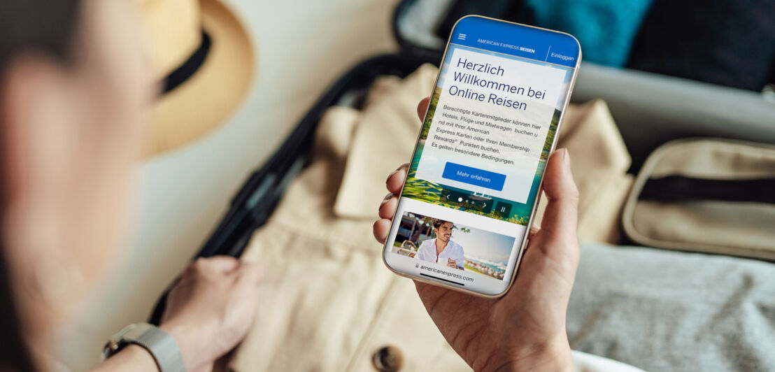 Eine Person hält ein Smartphone mit dem Online-Reiseservice von American Express auf dem Display über einen gepackten Koffer.