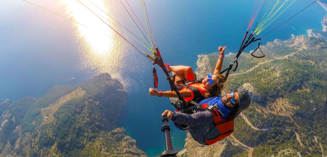 Aufsicht zweier Personen während eines Tandemflugs beim Paragliding über einer Küstenregion.