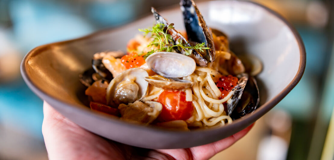 Eine Person hält eine Schale mit Pasta, die mit verschiedenen Muschelsorten und Tomaten angerichtet ist.