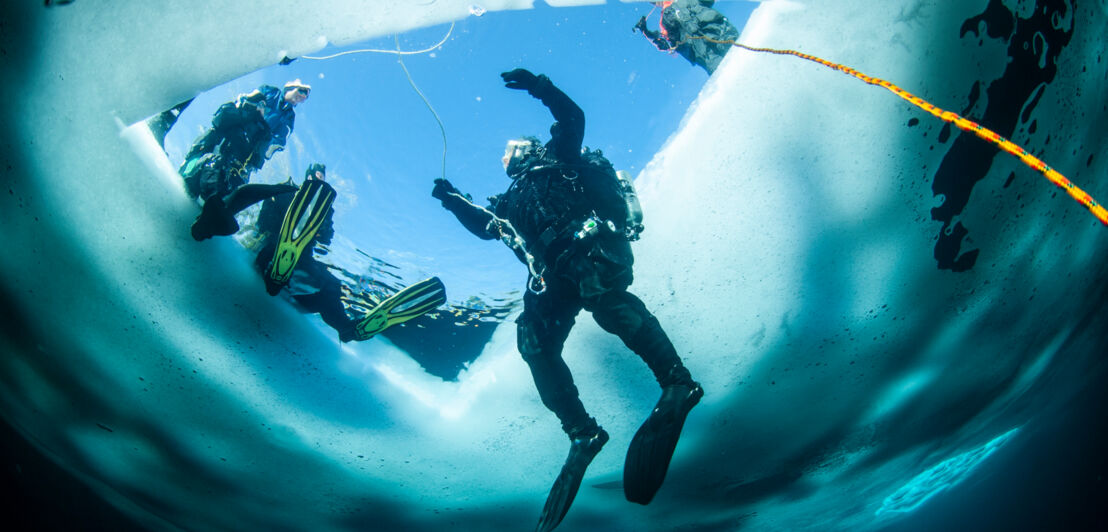 Eine Person in Taucherausrüstung, die aus dem zugefrorenen Wasser aus einem Loch in der Eisdecke auftaucht. Am Rand sitzt eine weitere Person.