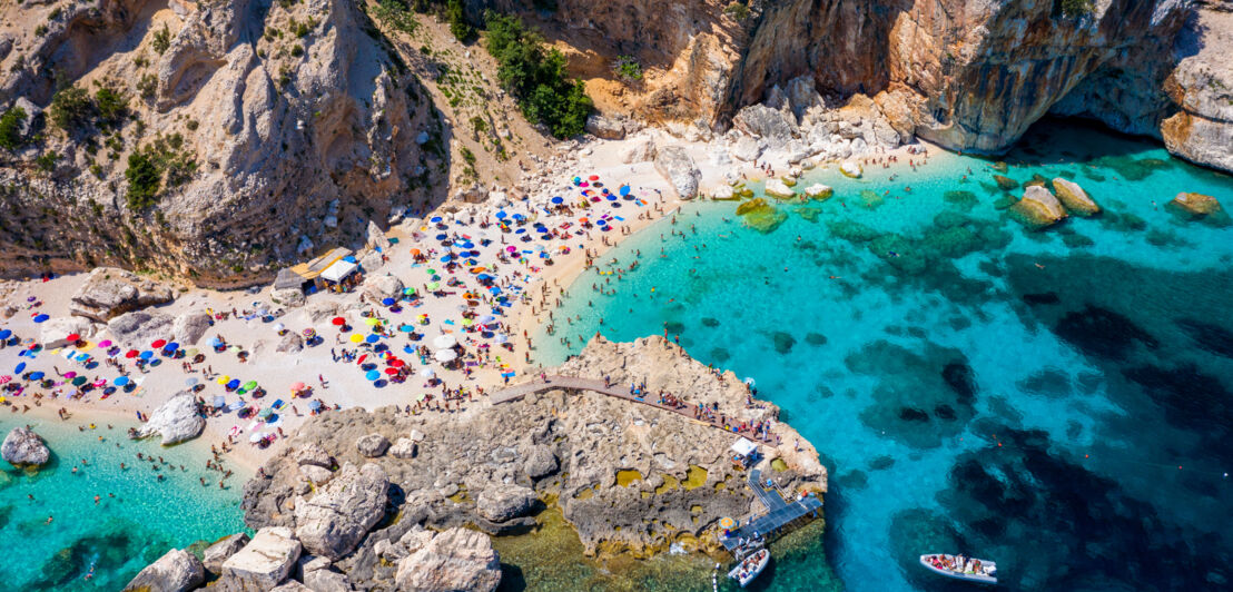 Aufsicht einer kleinen Felsenbucht mit belebtem Sandstrand und türkisblauem Wasser.