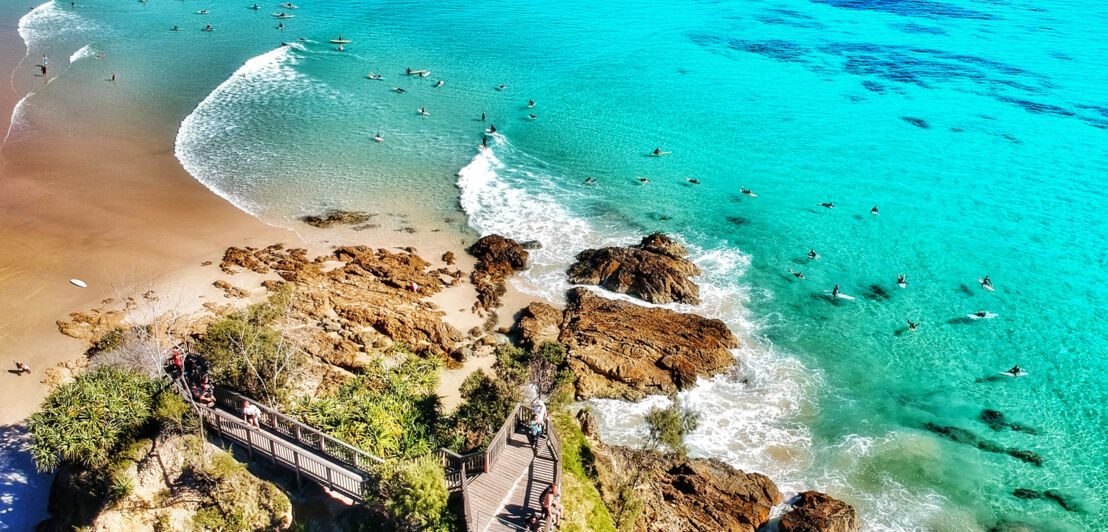 Aufsicht eines Küstenabschnittes mit Felsen und Sandstrand, dahinter zahlreiche Surfende im türkisblauen Wasser.