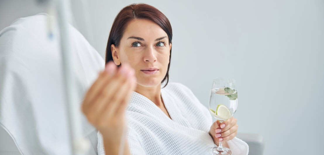 Frau in einer Schönheitsklinik reguliert ihre Vitamininfusion und hält ein Glas Wasser in der Hand.