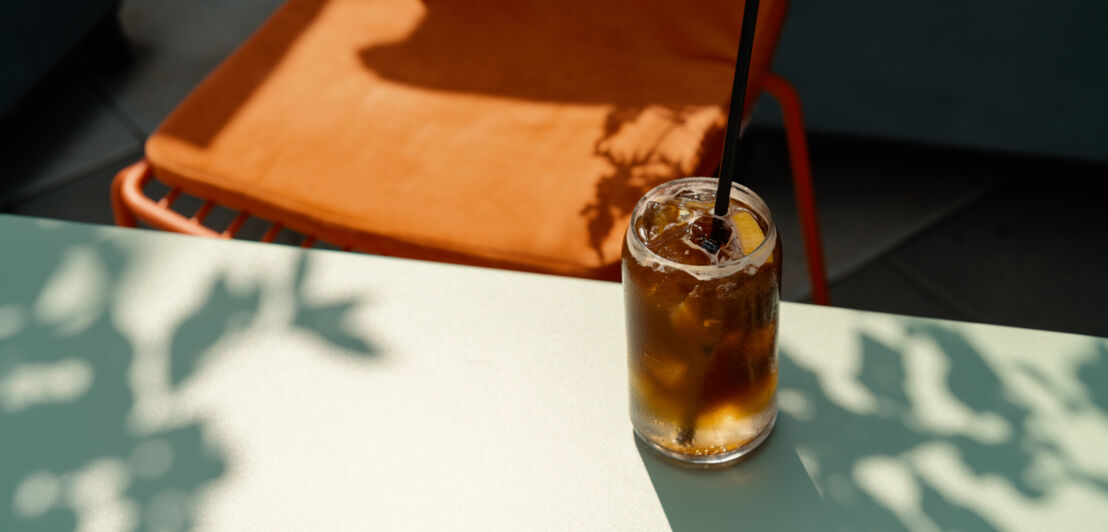 Ein Glas Espresso Tonic mit Strohhalm auf einem Tisch, dahinter steht ein orangefarbener Stuhl.