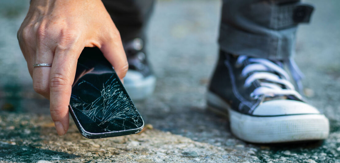 Eine Hand greift nach einem Smartphone mit zersprungenem Display am Asphaltboden.