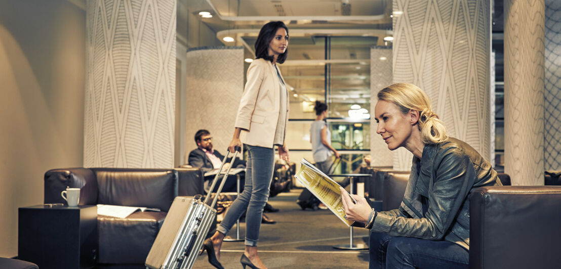 Eine Frau sitzt in einem Ledersessel und blättert in einem Magazin, im Hintergrund sind weitere Menschen zu sehen