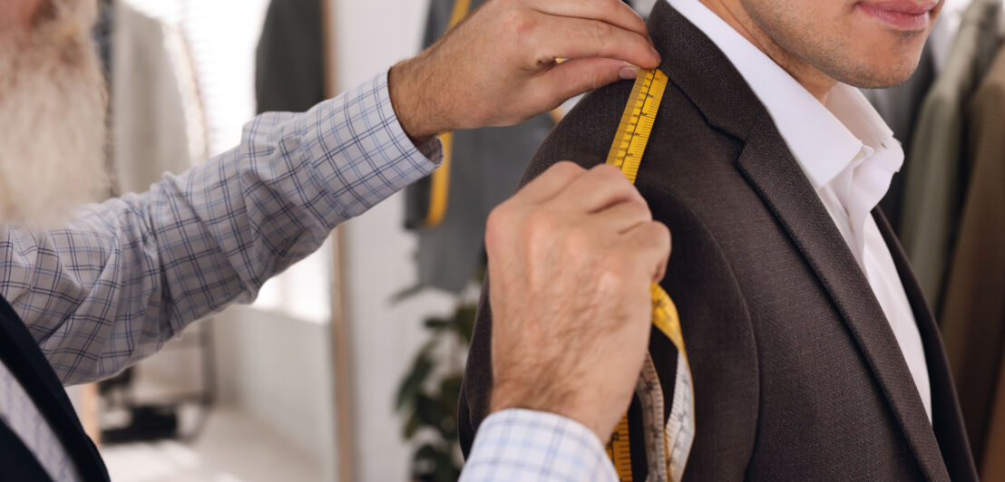 Ein Schneider misst in seinem Atelier die Schulterlänge eines von einem Kunden getragenen Jacketts, Nahaufnahme.