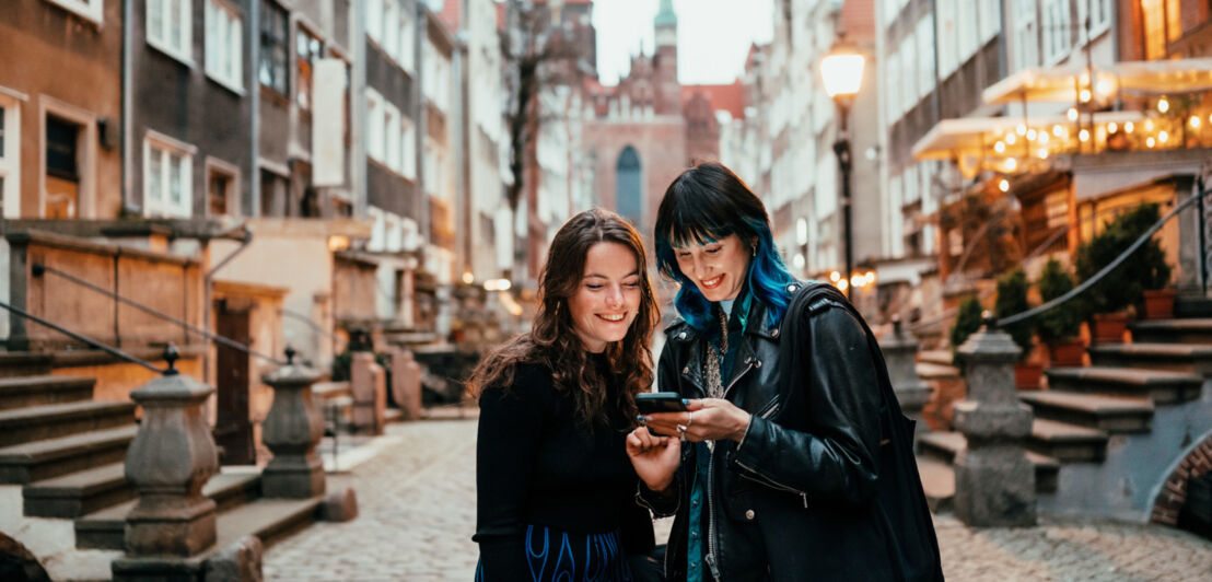 Zwei junge Frauen stehen in einer kopfsteingepflasterten Straße und blicken auf ein Smartphone, das eine von ihnen in der Hand hält.