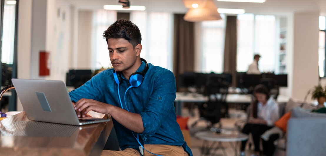 Geschäftsmann mit Kopfhörern sitzt in einem Coworking Space an einem Tisch und arbeitet am Laptop