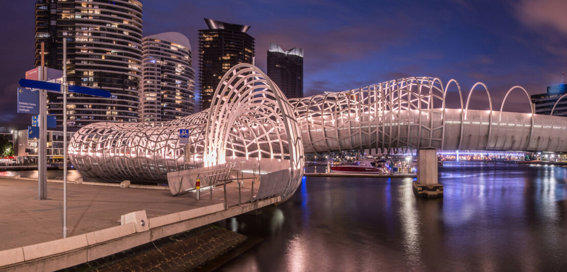 Geschwungene Brücke aus einer kunstvollen Stahlkonstruktion an der Promenade eines modernen Hafenviertels bei Nacht.