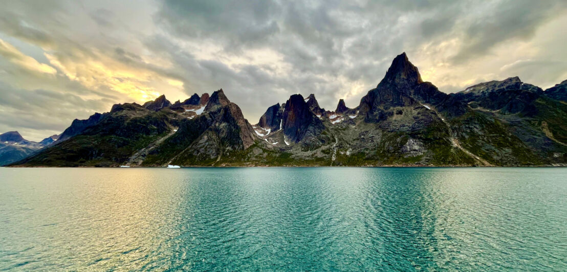 Das Bild zeigt hohe und spitze Berge. Im Vordergrund ist das grünlich schimmernde Wasser des Prins Christian Sunds zu sehen.