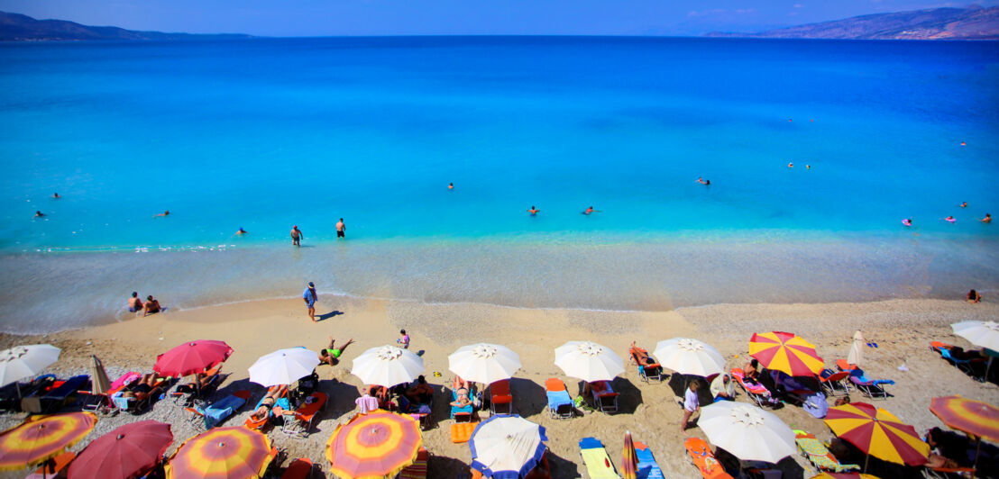 Aufsicht eines Sandstrandes mit Liegen und Sonnenschirmen am Meer mit Wasser von starker blauer Farbintensivität.