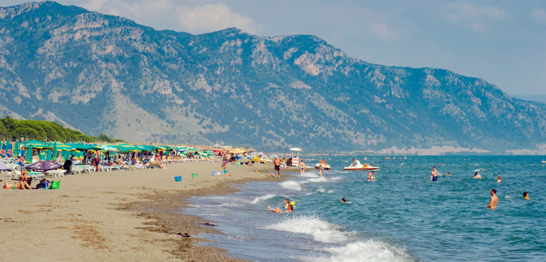 Sandstrand mit Personen in und am Wasser vor Bergpanorama.