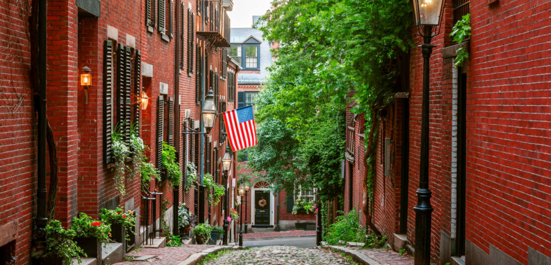 Acorn Street in Boston mit Kopfsteinpflaster und Laternen, an einer Hauswand hängt eine USA-Flagge.