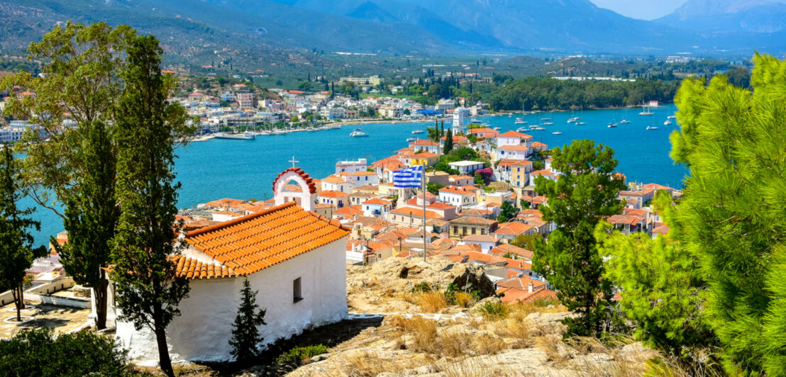 Blick auf einen mediterranen Ort von einem Felsen mit griechischer Flagge vor Küstenpanorama.