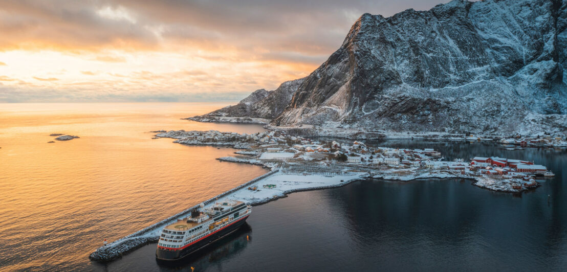 Luftaufnahme von einem Kreuzfahrtschiff, vor Anker an einer schneebedeckten Ortschaft vor kargem Bergpanorama bei Sonnenuntergang.