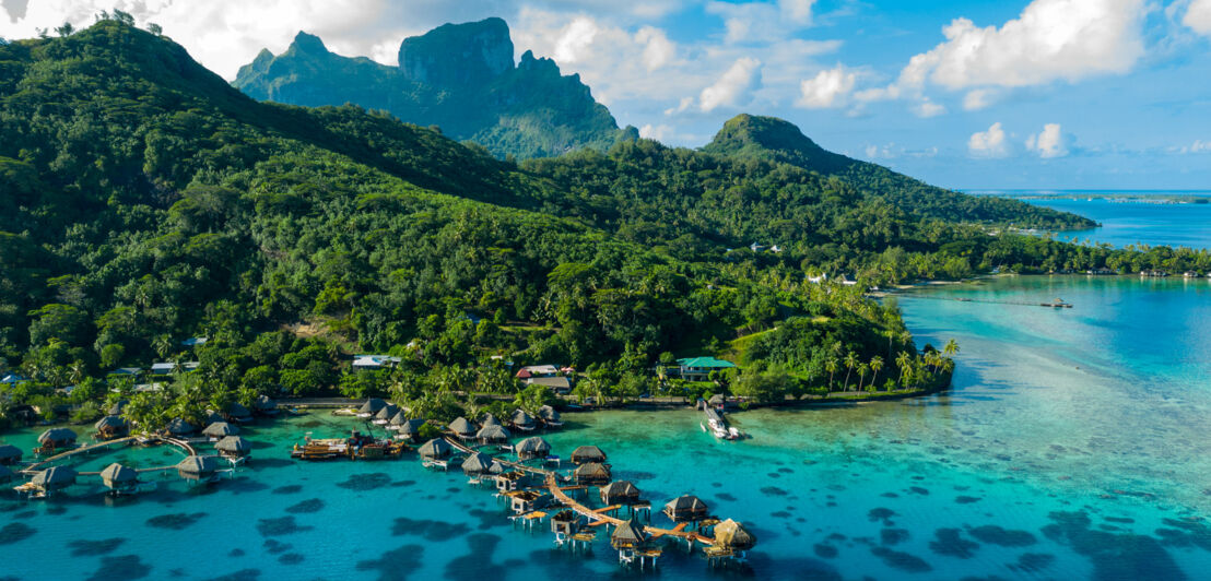 Vulkaninsel mit üppig grüner Vegetation, im Vordergrund Resort mit Bungalows auf Stelzen im türkisblauen Wasser.