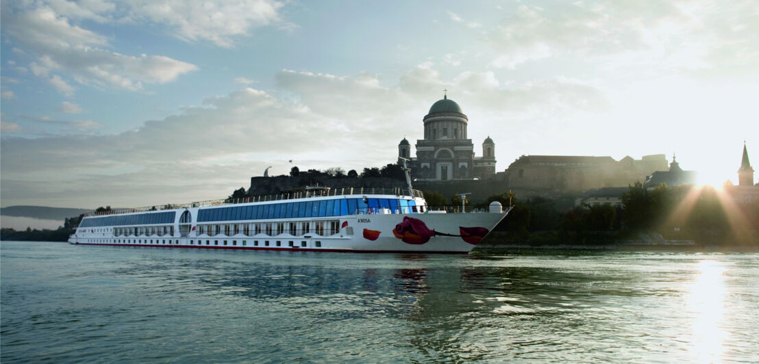 Ein Kreuzfahrtschiff von A-ROSA auf einem Fluss vor dem Panorama einer Stadt mit Kirche.