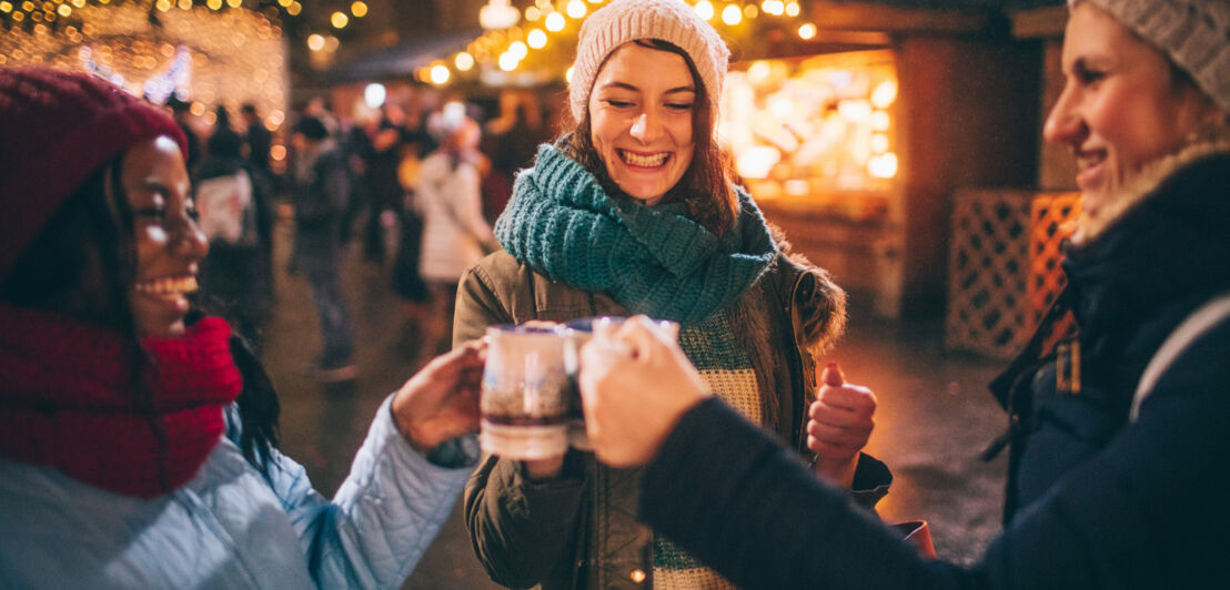 Drei Frauen in Winterkleidung stoßen mit Tassen auf einem Weihnachtsmarkt am Abend an.