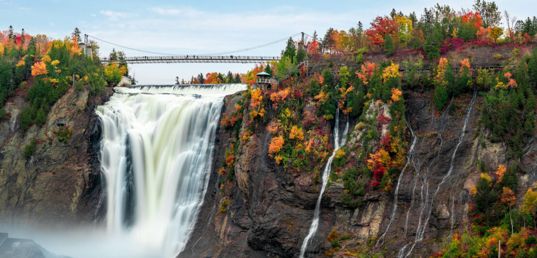 Personen auf einer Brücke über einem Wasserfall, umgeben von Bäumen mit herbstlicher Verfärbung.