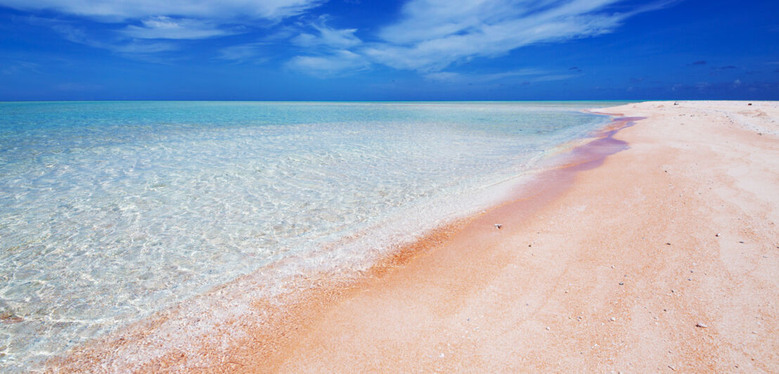 Weiter Sandstrand mit rosa Färbung an klarem Wasser unter blauem Himmel.