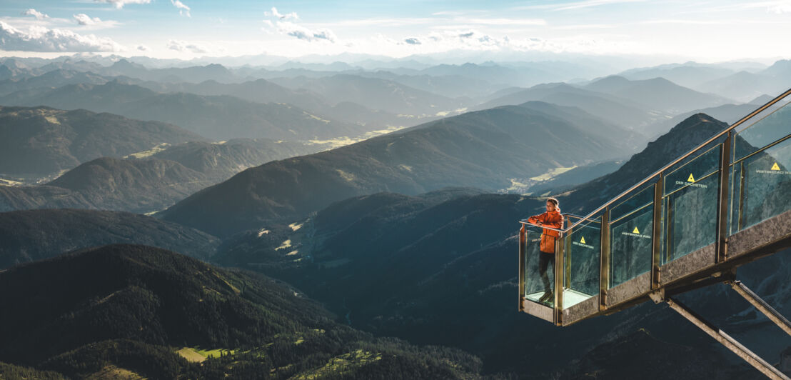 Eine Person steht auf einer gläsernen Aussichtsplattform vor Berglandschaft.