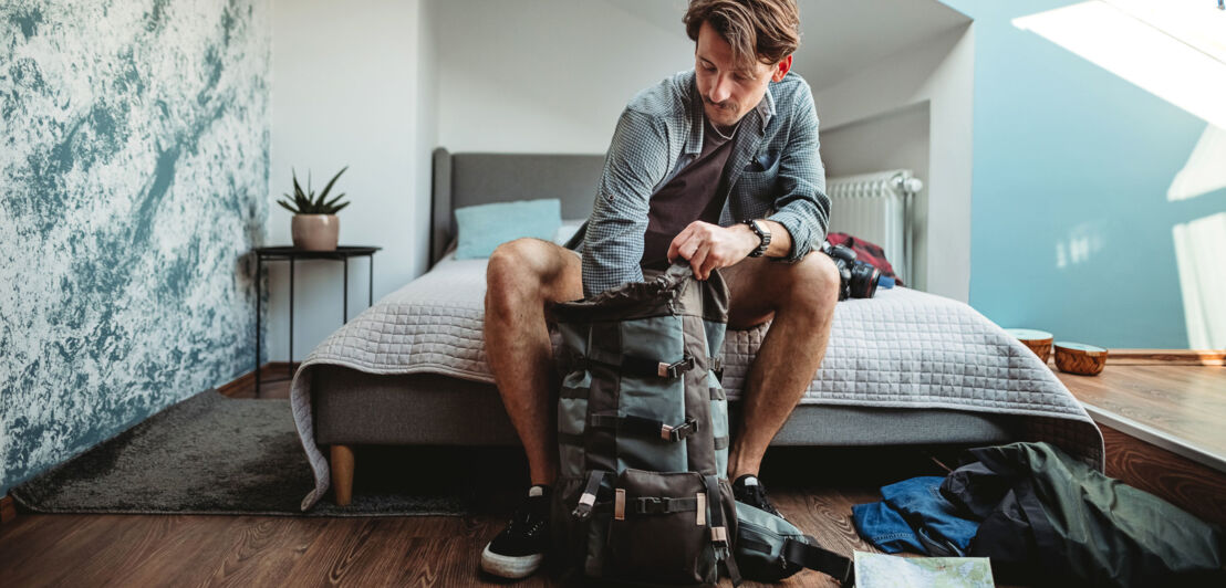 Ein Mann sitzt auf einem Bett und packt einen großen Rucksack.