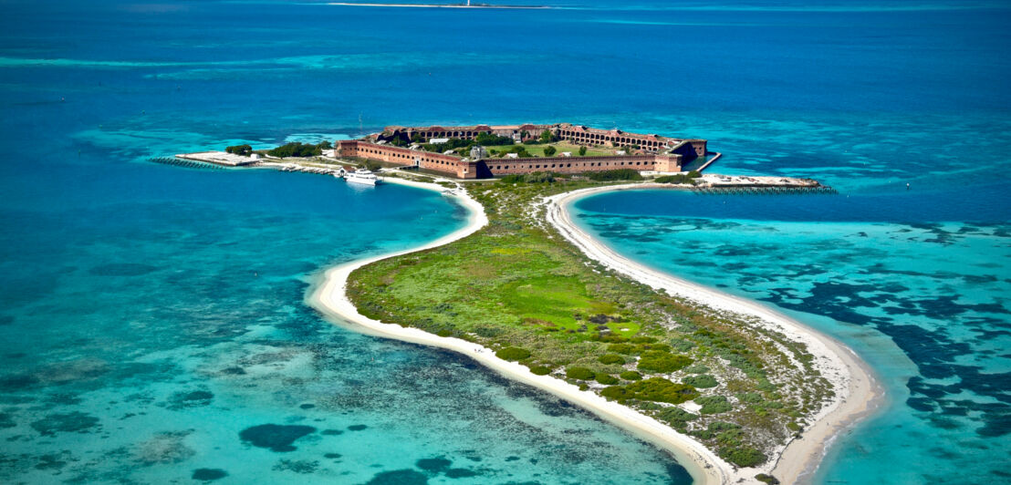 Eine Festung am Ende einer kleinen Insel mit Sandstrand im flachen, türkisblauen Meer.