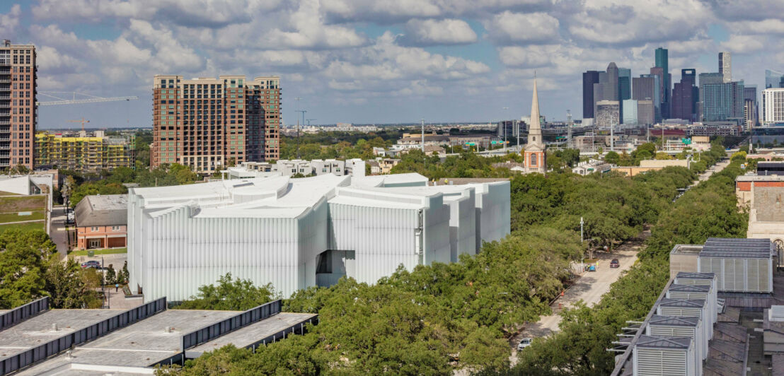 Ein modernes, weißes Museumsgebäude in einem Park in einer Stadt vor einer Skyline.