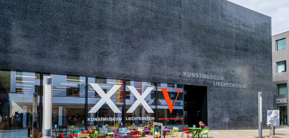 Außenansicht eines modernen, kubistischen Museumsgebäude aus schwarzem Basaltstein mit der Aufschrift Kunstmuseum Liechtenstein, im Vordergrund bunte Stühle vor einer Fensterfront.