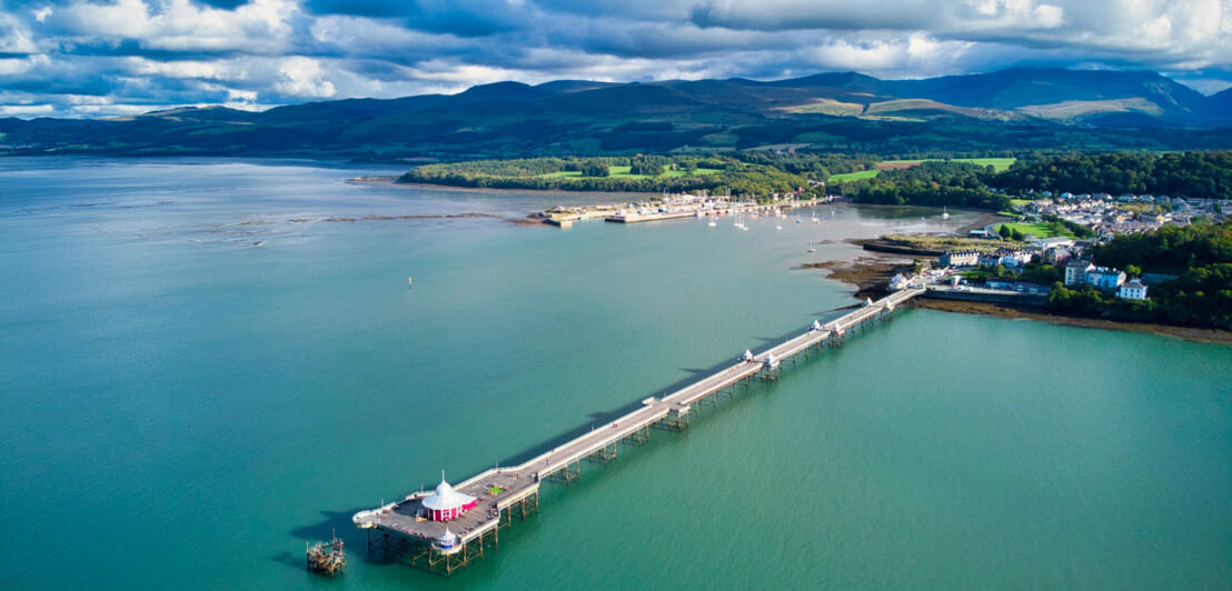 Langer Pier an einem Küstenort, im Hintergrund grüne Hügellandschaft.