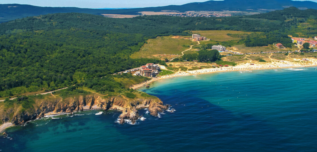 Küstenpanorama mit grüner Vegetation und vereinzelten Gebäuden an einem Sandstrand aus der Luftperspektive.