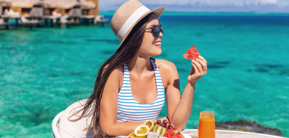 Eine Frau mit Sonnenhut sitzt an einem Tisch mit Obstteller und Fruchtsaft vor türkisblauem Meer mit Überwasser-Bungalows.