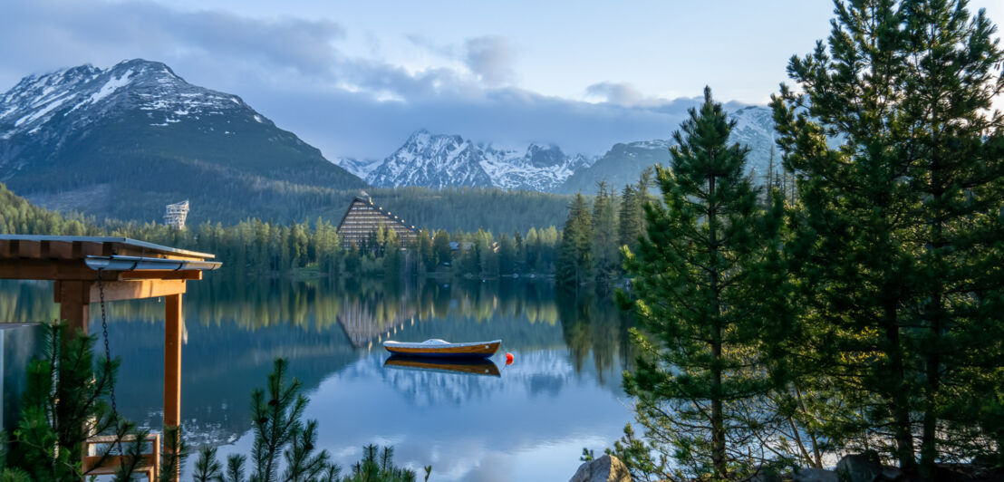 Ein idyllischer Bergsee mit Boot, umgeben von Nadelwald in der Abenddämmerung, im Hintergrund schneebedecktes Gipfelpanorama.