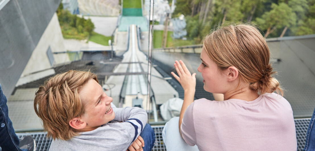 Zwei Kinder im Profil an einer Glasscheibe auf einer Aussichtsplattform einer Sprungschanze im Sommer.