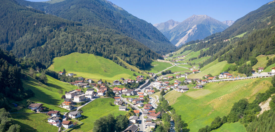 Ortschaft in einem grünen Tal mit Waldgebieten vor Bergpanorama.