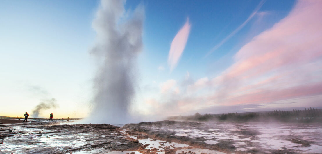 Ein sprudelnder Geysir auf einem Felsplateau unter blauem Himmel, im Hintergrund vereinzelte Personen.