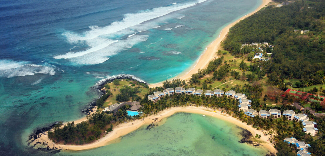 Luftaufnahme eines Küstenabschnitts mit Hotelresort inmitten tropischer Natur auf einer Landzunge mit Sandstrand, umgeben von türkisblauem Meer.