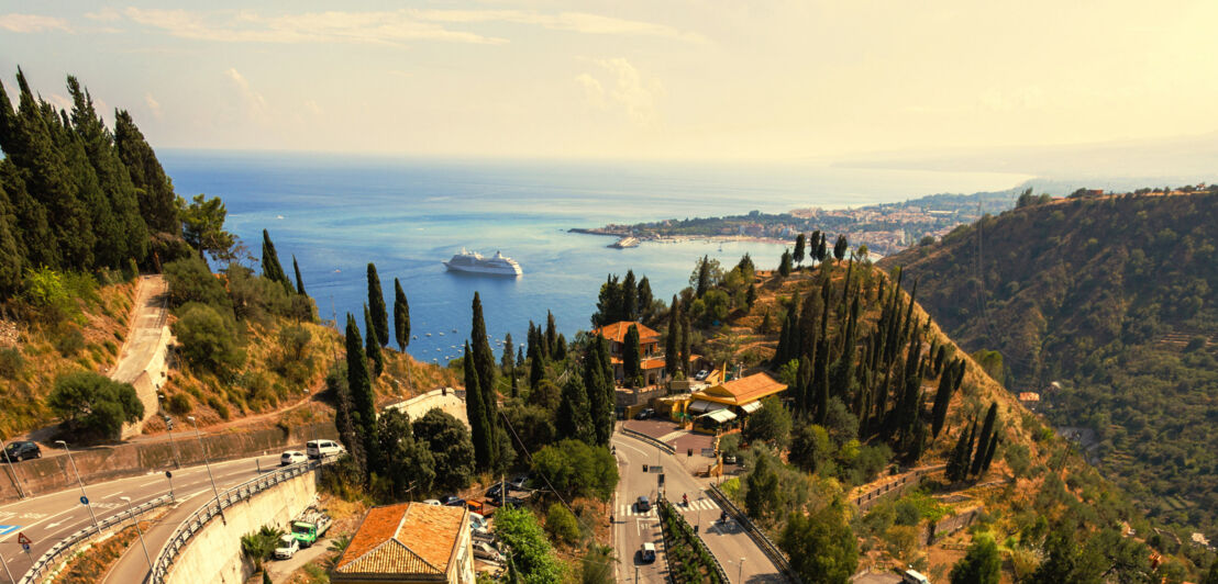 Serpentinenstraße in mediterraner Berglandschaft mit Zypressen mit Blick auf eine Meeresbucht mit Kreuzfahrtschiff.