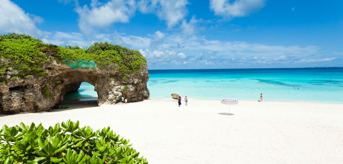 Personen an einem weißen Sandstrand mit bewachsenem Felsen an türkisblauem Meer.