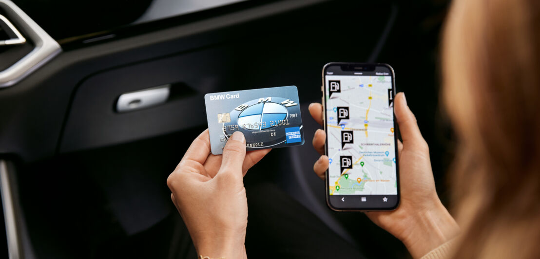 Eine BMW Card von American Express neben einem Smartphone mit Tankstellen-Icons auf einer Straßenkarte auf dem Display in den Händen einer Person in einem Auto.