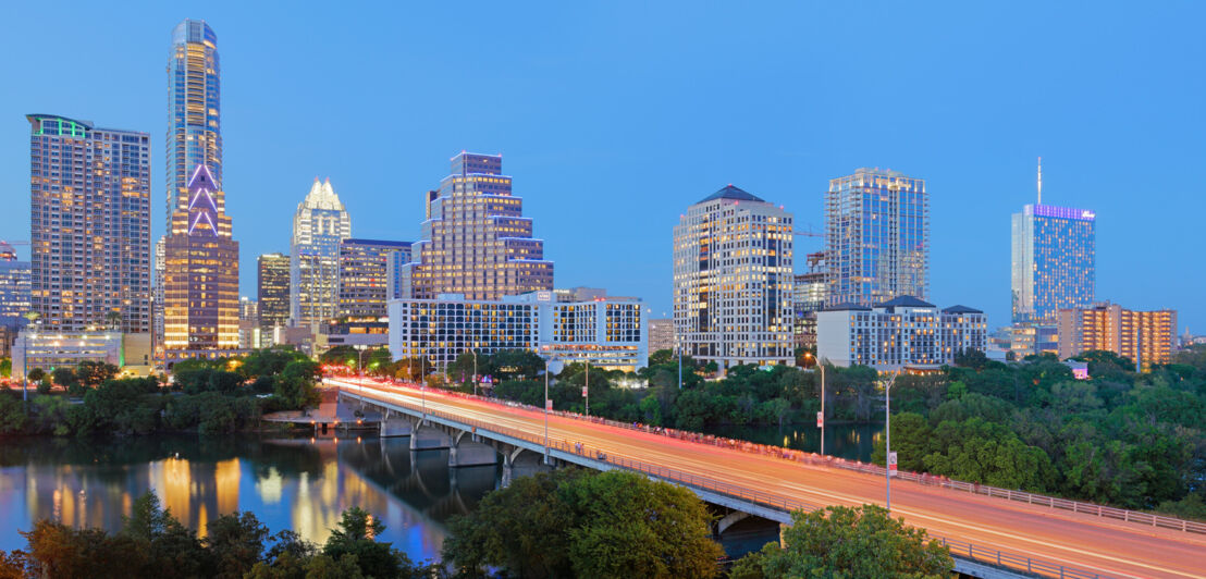 Beleuchtete Skyline von Austin mit Brücke über einem Fluss im Vordergrund in der Abenddämmerung.