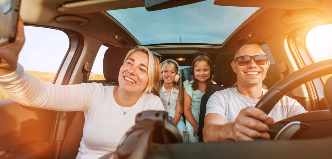 Eine Frau, ein Mann und zwei Mädchen, alle lächelnd, sitzen in einem Auto, die Frau auf dem Beifahrersitz macht ein Foto.