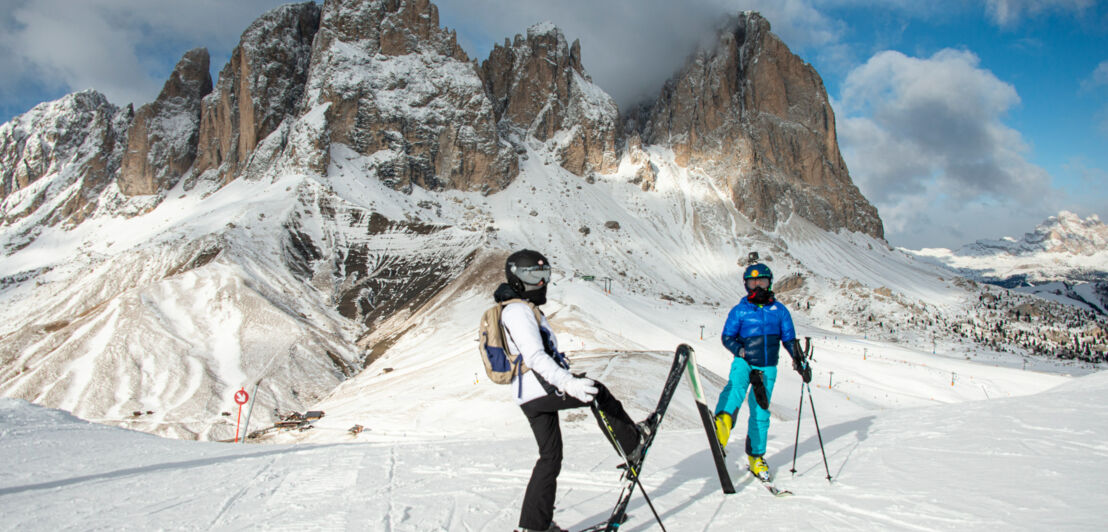 Zwei Personen stehen auf einer Skipiste vor einer Bergkulisse.