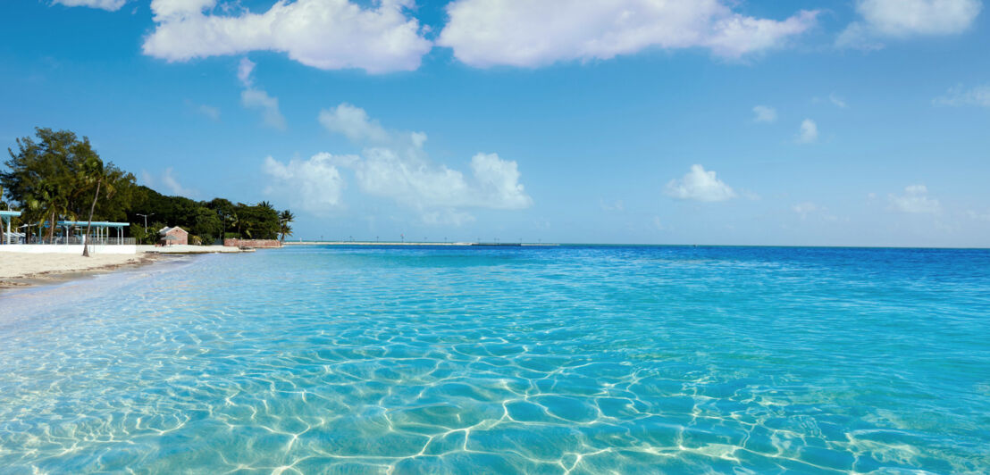 Flaches türkisblaues Meer vor einem weißen Sandstrand unter blauem Himmel.