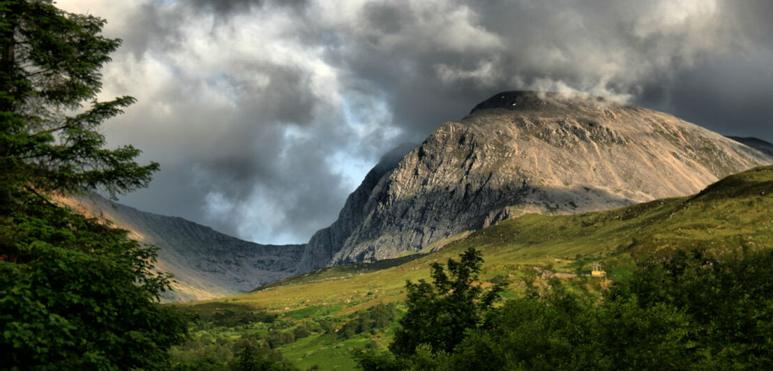 Blick auf den Berg Ben Nevis in Schottland bei stark bewölktem Himmel, im Vordergrund eine grüne Landschaft. 