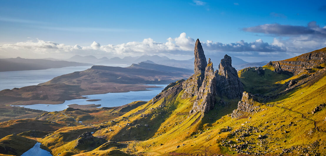 Blick auf den Old Man of Storr, ein markantes Felsmassiv, das von einer grünen Landschaft mit Seen umgeben ist. 